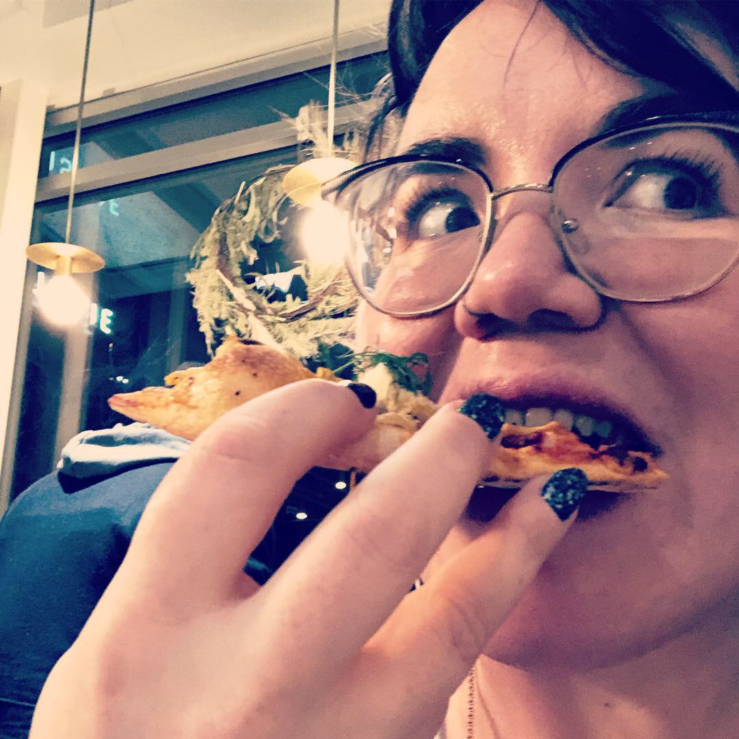 Close-up of Hannah biting into a slice of pizza, wide-eyed with joy 